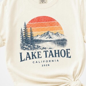 May include: Ivory-colored t-shirt with a graphic of a sunset over Lake Tahoe, California. The design includes mountains, trees, and the text "LAKE TAHOE CALIFORNIA 2026". The shirt is made of a soft material.