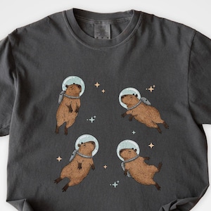 Cute capybara floating in space wearing an astronaut helmet and backpack. A quirky design for space lovers, capybara fans, and fans of kawaii and sci-fi aesthetic. Perfect funny animal gift or graphic tee for meme lovers, cottagecore fans, and rodent enthusiasts.