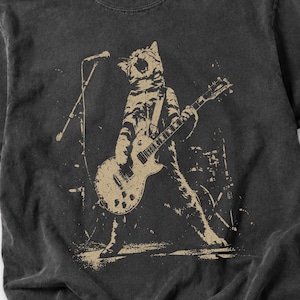 Grunge rock cat graphic featuring a roaring cat performing with an electric guitar on a dimly lit concert stage. A microphone stand, drum equipment, and distressed background details create the atmosphere of a live punk show. The vintage worn print style makes this design ideal for cat owners, musicians, guitar lovers, concertgoers, alternative fashion fans, and people who enjoy humorous animal band artwork.
