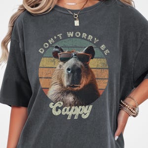 Woman wearing a pepper-colored t-shirt with a retro graphic of a capybara in sunglasses. The text reads 'Don't Worry Be Cappy.' Perfect for capybara fans, lovers of funny animal shirts, and vintage-style tees. A gift for those who enjoy animal memes.