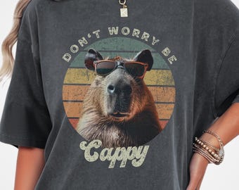 May include: Dark grey t-shirt featuring a capybara wearing sunglasses, set against a retro sunset design. The shirt includes the text "DON'T WORRY BE CAPPY" in a vintage font. The shirt is a relaxed fit.