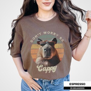 Woman wearing an espresso-colored t-shirt featuring a retro graphic of a capybara in sunglasses. The shirt says 'Don't Worry Be Cappy.' Perfect for capybara fans, vintage-style tee enthusiasts, and lovers of funny animal shirts. A cute gift idea.