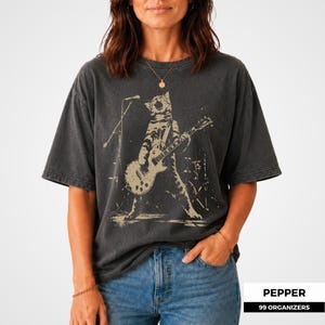 Rock cat artwork featuring a tabby cat in a powerful wide-legged stage pose while playing electric guitar and howling into a microphone. Distressed ink, concert lighting, and background drum equipment give the illustration a vintage band merchandise feel. The design combines feline humor with punk attitude, making it a memorable choice for cat dads, cat moms, musicians, music lovers, and alternative clothing fans.