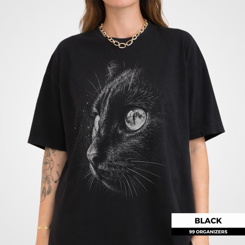 Celestial cat shirt featuring a dramatic close up illustration of a cat’s face with detailed fur texture and halftone shading. The cat’s eye contains a reflection of a full moon, creating a mystical and atmospheric effect. The high contrast monochrome ink style gives the design a vintage engraved look, making it an ideal graphic tee for cat lovers and fans of dark, artistic designs.