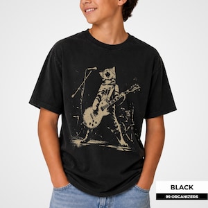 May include: A black short-sleeve t-shirt featuring a beige graphic print of a cat standing on its hind legs while playing an electric guitar in front of a microphone stand. The design has a distressed, textured appearance on the fabric.