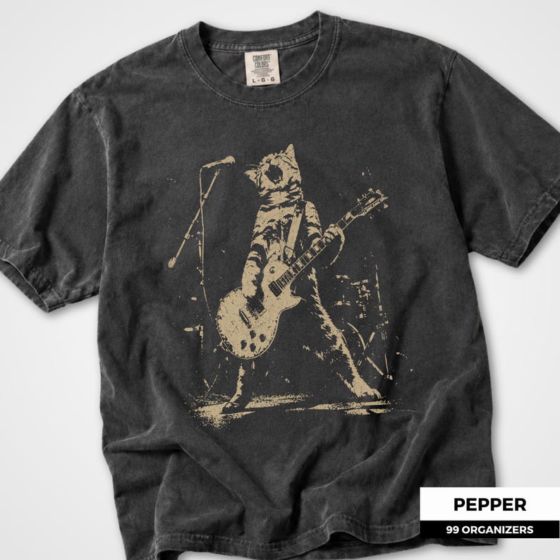 Grunge rock cat graphic featuring a roaring cat performing with an electric guitar on a dimly lit concert stage. A microphone stand, drum equipment, and distressed background details create the atmosphere of a live punk show. The vintage worn print style makes this design ideal for cat owners, musicians, guitar lovers, concertgoers, alternative fashion fans, and people who enjoy humorous animal band artwork.