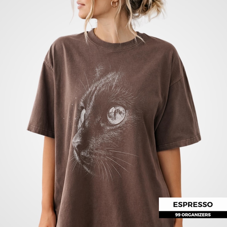 shading and engraved style lines. The cat’s eye reflects a full moon, creating a subtle celestial theme. The high contrast monochrome design gives it a vintage and mysterious aesthetic.