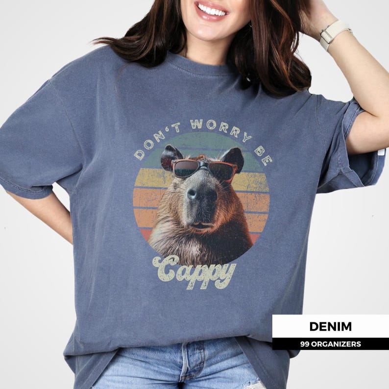 Woman wearing a denim-colored t-shirt with a retro graphic of a capybara in sunglasses. The text reads 'Don't Worry Be Cappy.' Ideal for capybara lovers, vintage-style enthusiasts, and fans of funny animal shirts. A cute and quirky gift idea