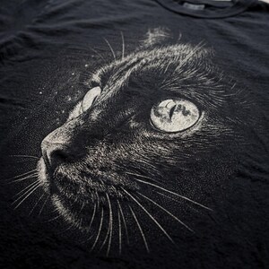 Vintage cat graphic tee featuring a detailed close up of a black cat’s face with intricate halftone shading and fine line texture. The cat’s eye reflects a full moon, adding a subtle celestial element. The monochrome distressed style creates a dark, moody aesthetic, making it a great shirt for cat lovers and fans of gothic or dark academia style.