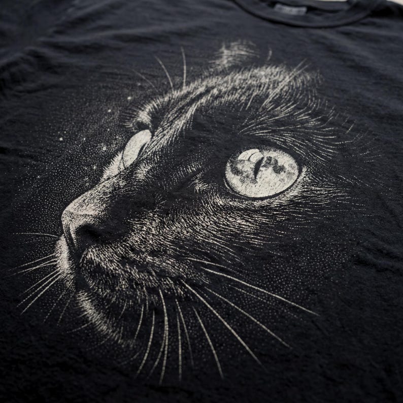Vintage cat graphic tee featuring a detailed close up of a black cat’s face with intricate halftone shading and fine line texture. The cat’s eye reflects a full moon, adding a subtle celestial element. The monochrome distressed style creates a dark, moody aesthetic, making it a great shirt for cat lovers and fans of gothic or dark academia style.