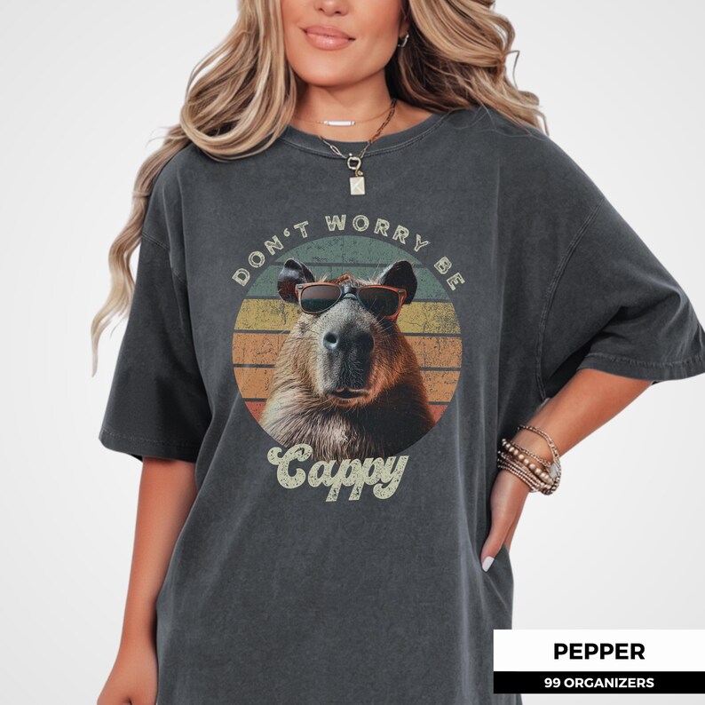 Woman wearing a pepper-colored t-shirt with a retro graphic of a capybara in sunglasses. The text reads 'Don't Worry Be Cappy.' Perfect for capybara fans, lovers of funny animal shirts, and vintage-style tees. A gift for those who enjoy animal memes.