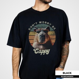 Puede incluir: Camiseta negra con un carpincho con gafas de sol, sobre un dise&ntilde;o retro de puesta de sol. El texto "DON'T WORRY BE CAPPY" y "Cappy" est&aacute;n impresos en la camiseta. Cuello redondo.