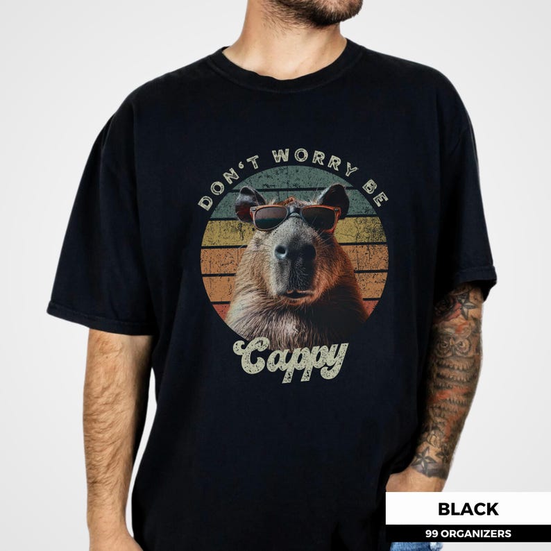 Puede incluir: Camiseta negra con un carpincho con gafas de sol, sobre un dise&ntilde;o retro de puesta de sol. El texto "DON'T WORRY BE CAPPY" y "Cappy" est&aacute;n impresos en la camiseta. Cuello redondo.