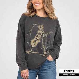 May include: A charcoal grey long-sleeve crewneck sweatshirt featuring a beige graphic print of a cat playing an electric guitar on stage with a microphone stand. The design has a distressed, weathered appearance on the fabric.
