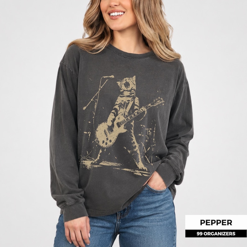 May include: A charcoal grey long-sleeve crewneck sweatshirt featuring a beige graphic print of a cat playing an electric guitar on stage with a microphone stand. The design has a distressed, weathered appearance on the fabric.