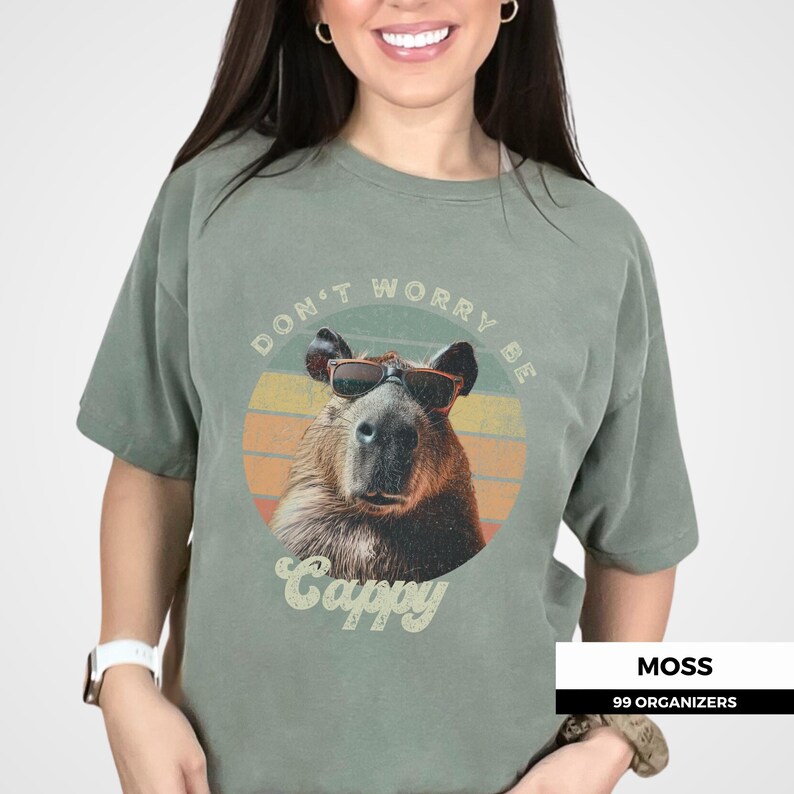 Woman wearing a moss green t-shirt featuring a retro-style capybara graphic with sunglasses. The shirt reads 'Don't Worry Be Cappy.' Ideal for capybara lovers, fans of funny animal shirts, and vintage-style tee enthusiasts. A cute and quirky gift.
