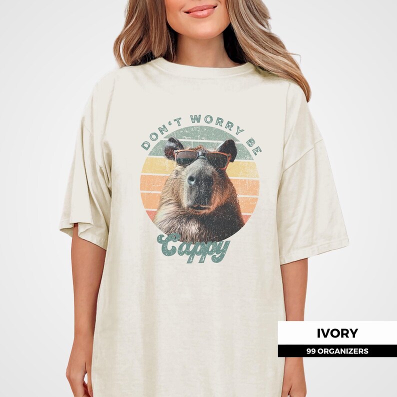 Woman wearing an ivory t-shirt with a retro graphic of a capybara in sunglasses. The text says 'Don't Worry Be Cappy.' Ideal for capybara fans, funny animal shirt lovers, and vintage tee enthusiasts. A cute gift for those who enjoy animal memes.