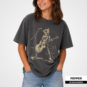May include: A charcoal gray short-sleeve t-shirt featuring a distressed beige graphic print of a cat playing an electric guitar on stage with a microphone stand. The shirt has a crew neckline and a relaxed fit, made of a washed-out cotton fabric.