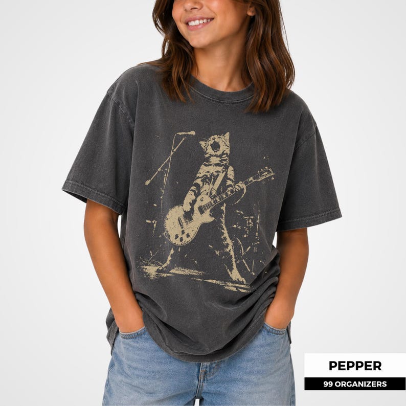 May include: A charcoal gray short-sleeve t-shirt featuring a distressed beige graphic print of a cat playing an electric guitar on stage with a microphone stand. The shirt has a crew neckline and a relaxed fit, made of a washed-out cotton fabric.