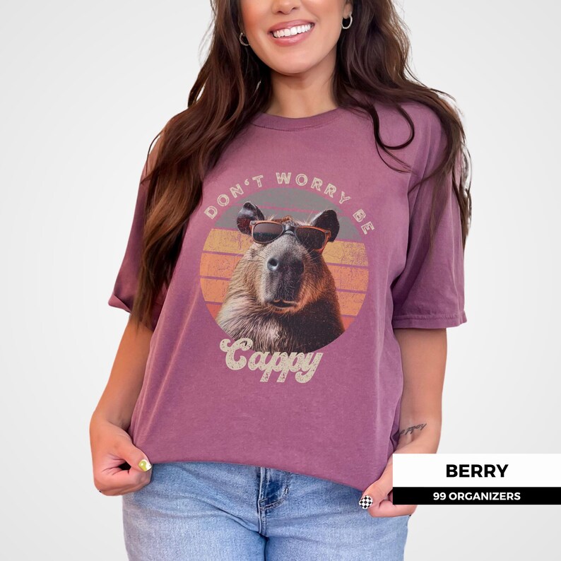 Woman wearing a berry-colored t-shirt featuring a retro graphic of a capybara in sunglasses. The shirt says 'Don't Worry Be Cappy.' Ideal for capybara lovers, vintage-style tee fans, and those who enjoy funny animal shirts. A cute and funny gift idea