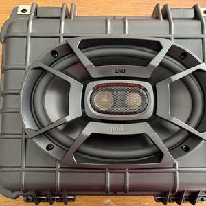 May include: A black oval Polk audio speaker mounted onto a dark gray, rectangular, hard-shell protective case with a grid-like exterior pattern. The speaker features a black grille, a central tweeter, and the text Dynamic Balance and Polk.