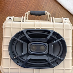 May include: A beige, rectangular speaker case with a black speaker grill. The speaker grill has an oval shape with the brand name "polk" visible. The case has a black handle and is on a wooden surface.