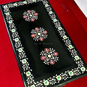 May include: A rectangular black table with floral inlay designs. The table features three central floral medallions and a border of white vines with green and yellow accents. The text "DECOR ENVOGUE" is visible.