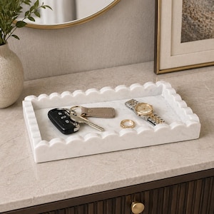 Op de afbeelding: Een rechthoekige witte marmeren tray met een geschulpte rand bevat sleutels, een horloge en een ring. De tray staat op een lichtgekleurd oppervlak, met een goudomrande spiegel en kunstwerk op de achtergrond.