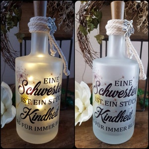 Könnte beinhalten: Eine gefrostete Glasflasche mit einem Seil und einem silbernen Anhänger. Die Flasche hat einen weißen Hintergrund mit einem schwarzen Design, auf dem "Eine Schwester ist ein Stück Kindheit" und "Für immer ble." steht. Die Flasche wird von innen mit weißen Lichtern beleuchtet.
