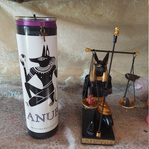 K&ouml;nnte beinhalten: Schwarzes und goldenes Anubis-Kerzen- und Statuenset. Die Kerze ist schwarz mit einem violetten Etikett und einer goldenen Anubis-Figur. Die Statue ist golden und schwarz und zeigt Anubis, der eine Waage h&auml;lt.