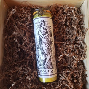 May include: A yellow candle with a black and white image of a figure holding a lyre. The candle has a gold sun charm on top and the text "APOLLO." printed on the bottom.