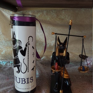 K&ouml;nnte beinhalten: Schwarze und goldene Anubis-Kerze mit einem violetten Band und einem goldenen Ankh-Anh&auml;nger. Die Kerze steht neben einer schwarzen und goldenen Anubis-Statue, die eine Waage h&auml;lt.