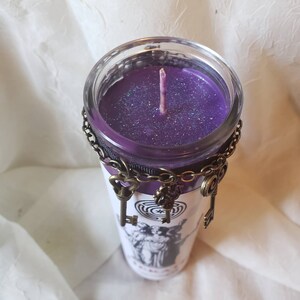 May include: A purple candle in a clear glass jar, adorned with a decorative chain and antique-style key charms. The jar features a black and white graphic design and a purple wick.