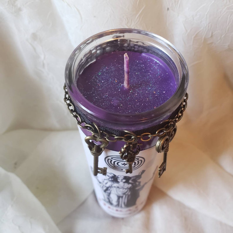 May include: A purple candle in a clear glass jar, adorned with a decorative chain and antique-style key charms. The jar features a black and white graphic design and a purple wick.