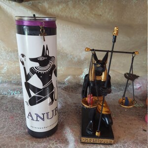 K&ouml;nnte beinhalten: Schwarzes und goldenes Anubis-Kerzen- und Statuenset. Die Kerze ist schwarz mit einem violetten Etikett und einer goldenen Anubis-Figur. Die Statue ist golden und schwarz und zeigt Anubis, der eine Waage h&auml;lt.
