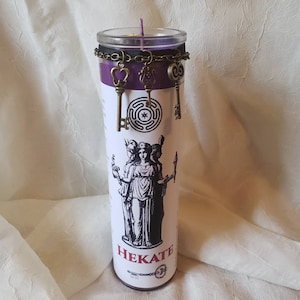 May include: A tall, cylindrical candle with a purple top and a clear glass container. The candle features a black and white illustration of a goddess and the word "HEKATE". Decorative bronze-colored key charms hang from a chain around the candle.
