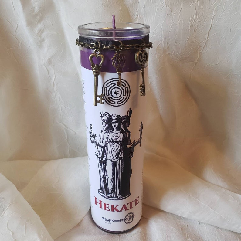May include: A tall, cylindrical candle with a purple top and a clear glass container. The candle features a black and white illustration of a goddess and the word "HEKATE". Decorative bronze-colored key charms hang from a chain around the candle.