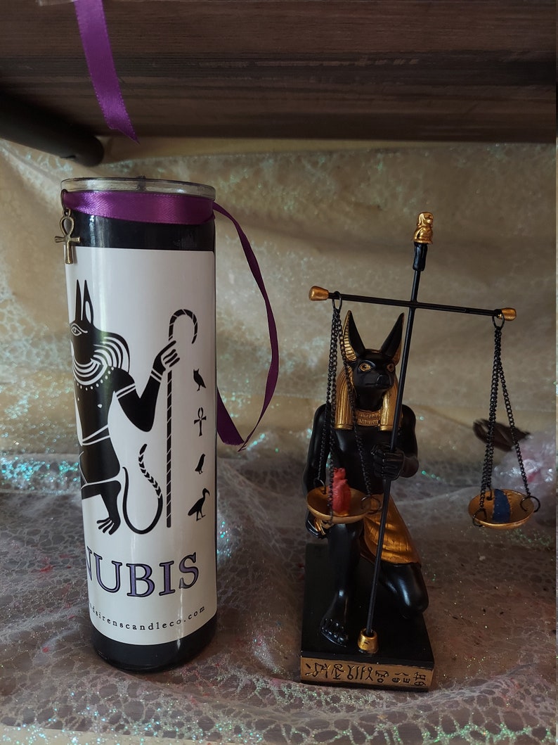 K&ouml;nnte beinhalten: Schwarze und goldene Anubis-Kerze mit einem violetten Band und einem goldenen Ankh-Anh&auml;nger. Die Kerze steht neben einer schwarzen und goldenen Anubis-Statue, die eine Waage h&auml;lt.