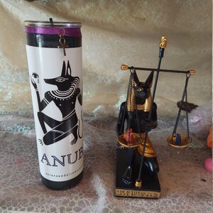 K&ouml;nnte beinhalten: Schwarze und goldene Anubis-Kerze und -Statue. Die Kerze ist schwarz mit einem violetten Band und einem goldenen Ankh-Anh&auml;nger. Die Statue zeigt Anubis, der eine Waage h&auml;lt. Die Statue steht auf einem schwarzen Sockel mit Hieroglyphen.