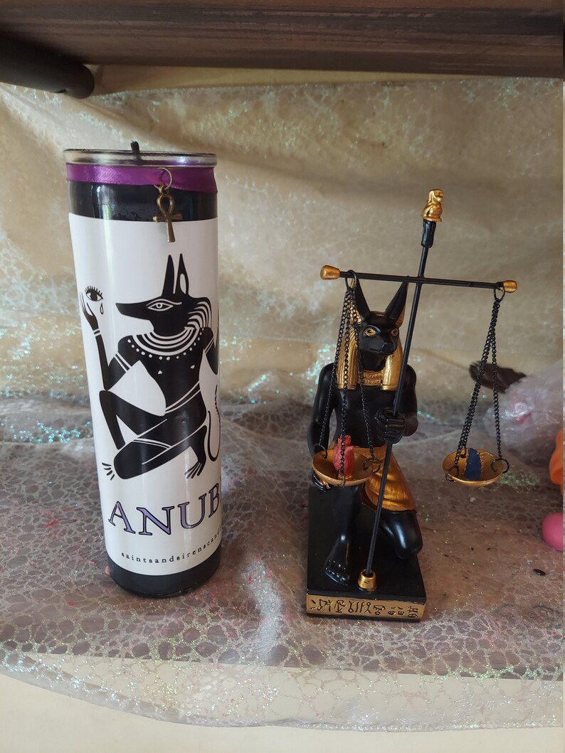 K&ouml;nnte beinhalten: Schwarze und goldene Anubis-Kerze und -Statue. Die Kerze ist schwarz mit einem violetten Band und einem goldenen Ankh-Anh&auml;nger. Die Statue zeigt Anubis, der eine Waage h&auml;lt. Die Statue steht auf einem schwarzen Sockel mit Hieroglyphen.