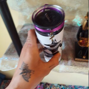 K&ouml;nnte beinhalten: Eine schwarze Kerze mit einem violetten Band und einem silbernen Anh&auml;nger in Form eines Kreuzes. Die Kerze befindet sich in einem Glasgef&auml;&szlig; mit einem wei&szlig;en Etikett, auf dem sich ein schwarz-wei&szlig;es Bild eines Vogels befindet.