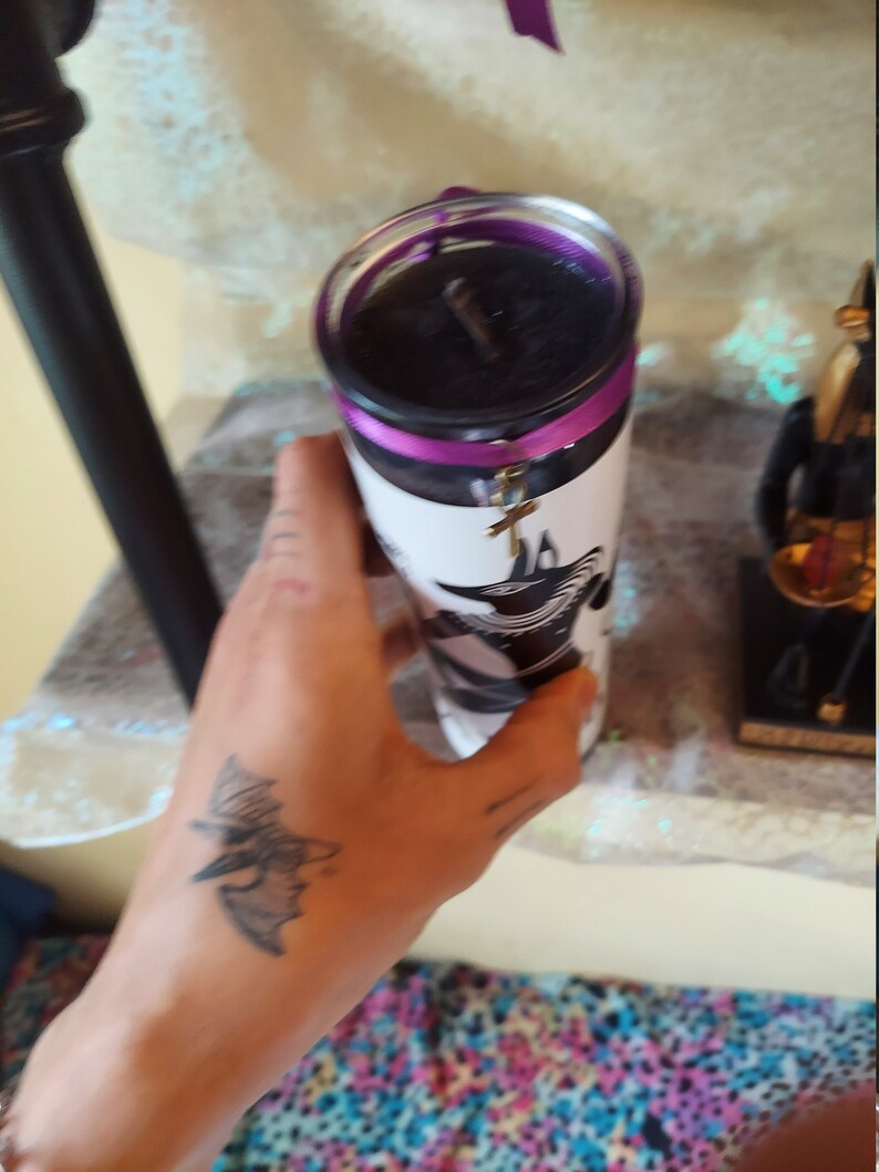 K&ouml;nnte beinhalten: Eine schwarze Kerze mit einem violetten Band und einem silbernen Anh&auml;nger in Form eines Kreuzes. Die Kerze befindet sich in einem Glasgef&auml;&szlig; mit einem wei&szlig;en Etikett, auf dem sich ein schwarz-wei&szlig;es Bild eines Vogels befindet.