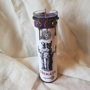May include: A tall, cylindrical candle with a purple wax top and a clear glass container. The candle features a black and white illustration of a goddess and the word "HEKATE". Decorative bronze-colored keys hang from a chain around the candle.