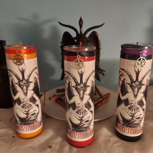 May include: Three tall, cylindrical candles with a Baphomet illustration and the word "Epitome" printed on them. Each candle has a different color scheme: orange and yellow, red and black, and purple and black. A pentagram charm hangs from each candle.