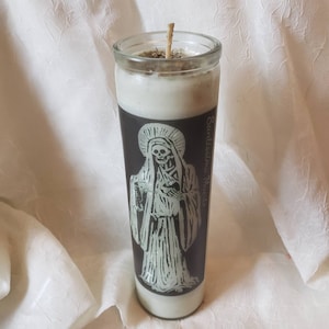 May include: A white candle with a black and white image of Santa Muerte, a skeletal figure in a robe, on the side. The candle is in a glass jar with a white wick.