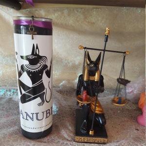 K&ouml;nnte beinhalten: Schwarze und goldene Anubis-Kerze mit einer schwarzen und goldenen Anubis-Statue mit Waagen. Die Kerze tr&auml;gt den Text "Anubis" und "Saints and Sirens Candles". Die Statue hat Hieroglyphen auf der Basis.