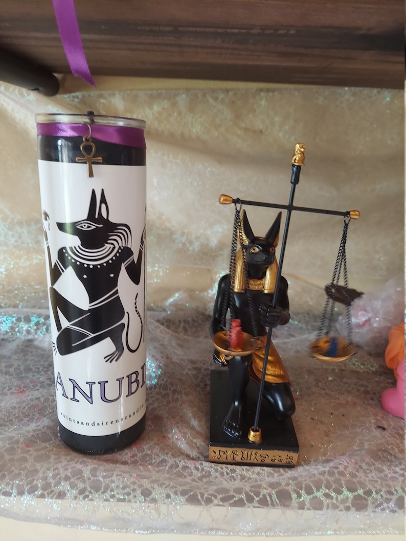 K&ouml;nnte beinhalten: Schwarze und goldene Anubis-Kerze mit einer schwarzen und goldenen Anubis-Statue mit Waagen. Die Kerze tr&auml;gt den Text "Anubis" und "Saints and Sirens Candles". Die Statue hat Hieroglyphen auf der Basis.