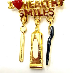 May include: Gold-tone pin with a red heart and the words "I ♥ HEALTHY SMILES".  The pin has three dangling charms: a toothbrush, a tube of toothpaste, and a dental pick.