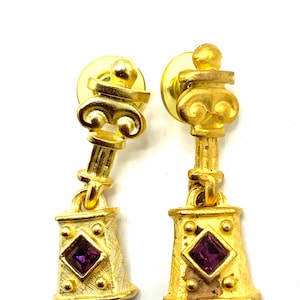 Pode incluir: Brincos de ouro com uma gema roxa em uma configuração quadrada. Os brincos apresentam um design de coluna de ouro com uma parte superior decorativa.