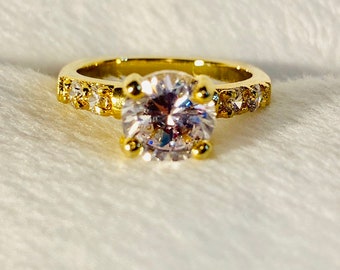 May include: A gold ring featuring a large, clear, round gemstone encircled by smaller, clear gemstones. The ring is displayed on a white, fluffy surface.